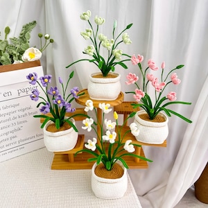 May include: A collection of handmade crochet flowers in white pots, arranged on a wooden tiered stand. The flowers are in various colors, including white, pink, purple, and yellow. The background includes a white curtain and a paper bag with French text.