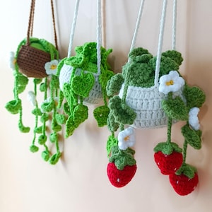 Puede incluir: Tres macetas colgantes de ganchillo con follaje verde y flores blancas. Una maceta es marrón, las otras son blancas. La tercera maceta tiene acentos de fresas rojas. Cada maceta cuelga de un cordón blanco.