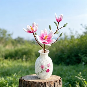 May include: A white crocheted vase holds a bouquet of handmade magnolia flowers. The flowers have pink and white petals with yellow centers. The vase is decorated with pink embroidered flowers and sits on a tree stump against a green background.