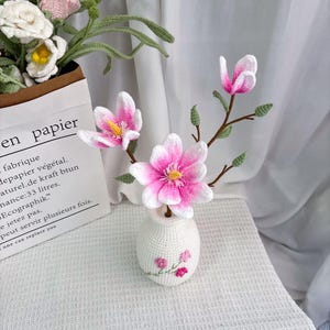 May include: A crocheted arrangement featuring pink and white magnolia flowers in a small white vase. The vase has embroidered pink flowers. The arrangement is on a white surface. The text "en papier" is visible on a paper bag in the background.