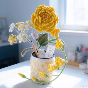 May include: A handmade crochet flower arrangement featuring a large yellow flower, smaller white and yellow flowers, and green leaves. The arrangement is in a yellow and white textured vase with a pencil and small cards. The cards have the word "hey" on them.