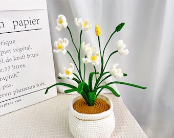 May include: A handcrafted crochet flower arrangement in a white pot. The arrangement features white flowers with yellow centers, green stems and leaves, and a yellow bud. The pot is white with a brown interior.