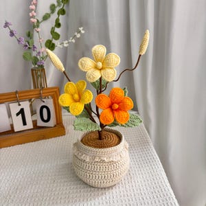 May include: A crocheted flower arrangement in a white crocheted pot. The flowers are yellow, orange, and cream coloured. The arrangement is on a white table with a wooden calendar that says "10" in the background.