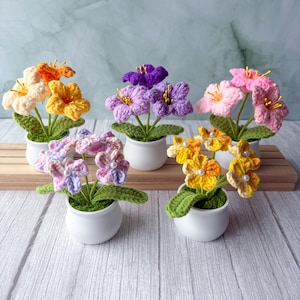 May include: A collection of handmade crochet flowers in small white pots. The flowers are in various colors, including orange, yellow, purple, and pink, with green leaves. The arrangement is displayed on a wooden surface, creating a charming, handcrafted aesthetic.