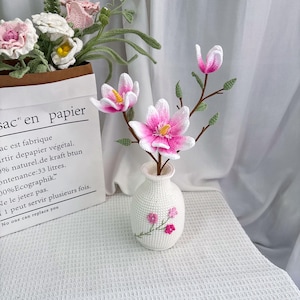 May include: A white crocheted vase holds pink and white crocheted magnolia flowers with yellow centers. The vase has a floral design. A paper bag with text and crocheted flowers is in the background.