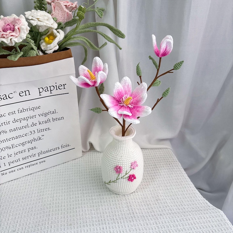 May include: A white crocheted vase holds pink and white crocheted magnolia flowers with yellow centers. The vase has a floral design. A paper bag with text and crocheted flowers is in the background.