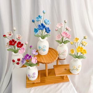 Op de afbeelding: Vijf gehaakte bloemstukken in witte keramische vazen, tentoongesteld op een houten standaard met meerdere niveaus. De bloemen zijn in tinten blauw, roze, rood, paars en geel.