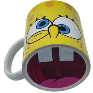May include: Yellow ceramic mug featuring a cartoon character's face with large blue eyes, a wide open mouth, and two large teeth. The mug has a white handle and is decorated with orange spots.