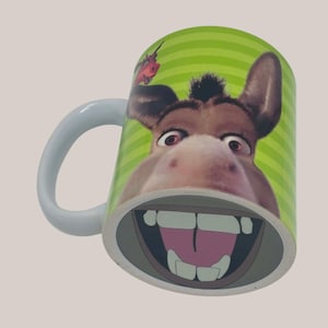 May include: White ceramic mug featuring a cartoon donkey face with a wide, toothy grin. The mug has a green striped background and a white handle. A red insect is on the side of the mug.