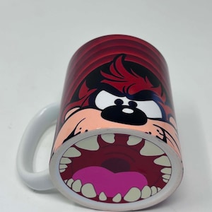 Può includere: Una tazza in ceramica bianca con una stampa grafica del personaggio del Diavolo della Tasmania. Il design mostra il volto del personaggio con una bocca spalancata, denti affilati e una lingua rosa su uno sfondo circolare rosso scuro.
