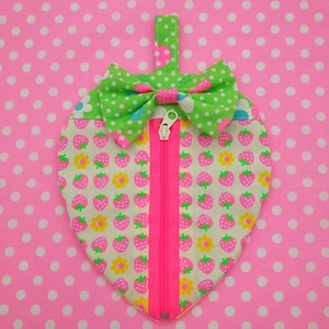 May include: A heart-shaped zippered pouch with a green and white polka dot bow. The pouch is made of a white fabric with a repeating pattern of pink strawberries and yellow flowers. The zipper is pink.