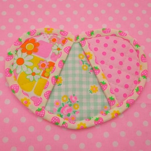 Può includere: Un sottopentola trapuntato a forma di cuore con un design patchwork. Il sottopentola è realizzato con tre tessuti diversi: un tessuto rosa con pois bianchi, un tessuto gingham verde e bianco con accenti floreali e un tessuto giallo e rosa con un motivo floreale. Il sottopentola è bordato con un tessuto bianco con stampa a fragola.