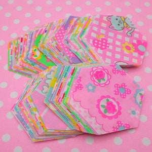 May include: A stack of pink fabric hexagons with various colorful patterns, including floral, polka dot, and geometric designs. The hexagons are arranged in a fan shape, with the largest hexagon at the bottom and the smallest hexagon at the top.