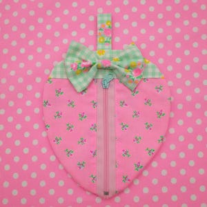 May include: A pink fabric strawberry-shaped pouch with a zipper closure and a green and white gingham bow. The pouch is decorated with a small floral pattern.