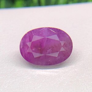 May include: An oval-cut gemstone with a vibrant magenta-pink hue. The stone features a faceted surface that creates a geometric pattern of light and shadow across its translucent body, highlighting its polished finish and clear internal structure.