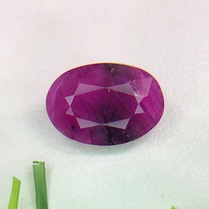 May include: An oval-shaped, faceted gemstone in a deep magenta-purple hue. The stone features a clear, symmetrical cut with visible internal inclusions and sharp, geometric facets that reflect light across its polished, translucent surface.