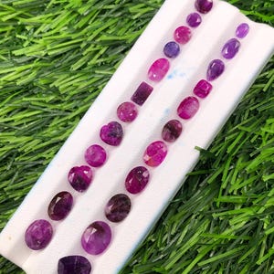 May include: A collection of faceted loose gemstones in various shades of purple, pink, and magenta, arranged in two parallel rows on a white display tray. The stones feature diverse shapes including oval, round, heart, and rectangular cuts.