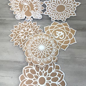 May include: Six white paper cutouts of mandalas and a peacock design on a brown wood background. The mandalas feature intricate patterns and the peacock design has a detailed body and tail feathers.