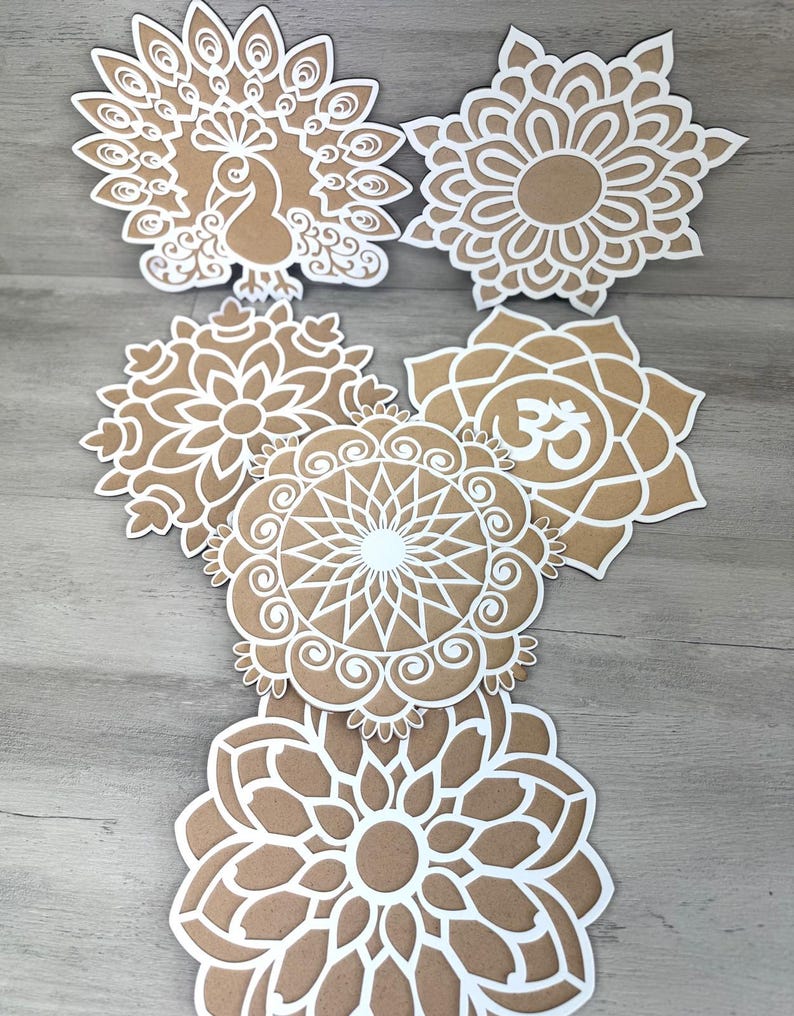 May include: Six white paper cutouts of mandalas and a peacock design on a brown wood background. The mandalas feature intricate patterns and the peacock design has a detailed body and tail feathers.