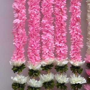 Op de afbeelding: Roze en witte bloemenslingers. De slingers zijn gemaakt van roze, gelaagde bloemblaadjes en witte lotusbloemen met groene bladeren. De slingers zijn aan elkaar geregen met een parelachtige draad.