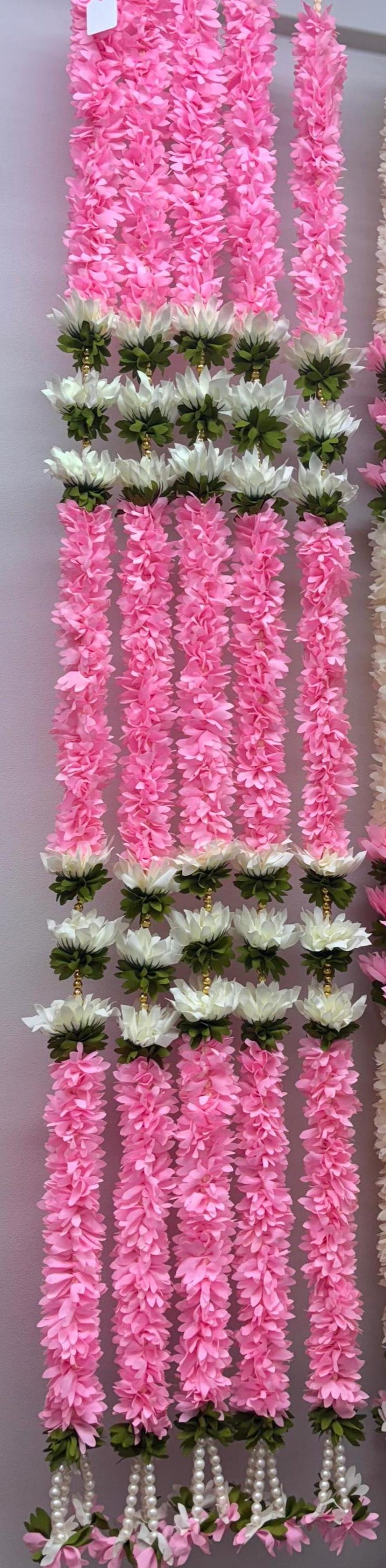Op de afbeelding: Roze en witte bloemenslingers. De slingers zijn gemaakt van roze, gelaagde bloemblaadjes en witte lotusbloemen met groene bladeren. De slingers zijn aan elkaar geregen met een parelachtige draad.