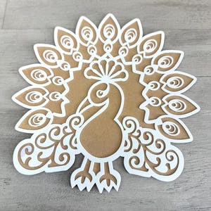 May include: A white paper cut-out of a peacock with a detailed design. The peacock is facing forward with its tail feathers spread out behind it.