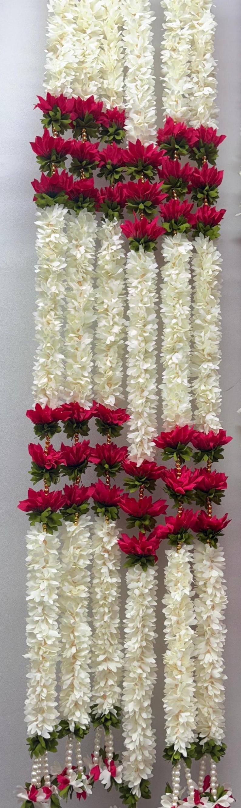 Op de afbeelding: Verticale afbeelding van verschillende strengen kunstmatige bloemenslingers. De slingers zijn voornamelijk wit met accenten van dieprode bloemen en groene bladeren. De achtergrond is effen wit.