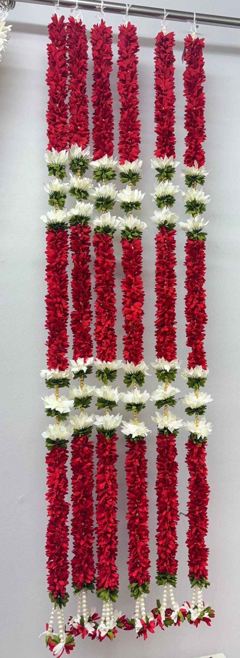Op de afbeelding: Verticale afbeelding van verschillende strengen rode bloemenslingers, afgewisseld met witte en groene bloemclusters. De slingers hangen tegen een lichtgrijze achtergrond, wat een levendige en feestelijke weergave cre&euml;ert. Elke slinger is ongeveer 122 cm lang.