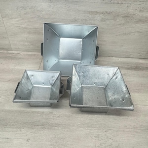 Könnte beinhalten: Drei Pflanzgefäße aus verzinktem Metall in verschiedenen Größen. Jedes Pflanzgefäß hat eine quadratische Form mit abgewinkelten Seiten und kleinen Griffen. Das Metall hat eine silberne Oberfläche und sichtbare Nieten. Die Pflanzgefäße sind ineinander verschachtelt.