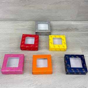 May include: A collection of colorful gift boxes with clear window tops. The boxes are in various colors, including red, yellow, pink, orange, and blue, each adorned with a gold paisley pattern. One box is silver.