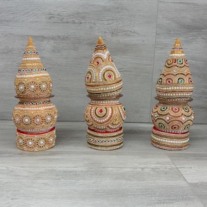 May include: Three decorative, ornate vessels with a gold and pearl beaded design. Each vessel has a bulbous base, a cylindrical midsection, and a conical top. Red, green, and white accents are incorporated. The vessels are arranged on a light grey surface.