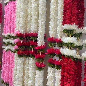 Op de afbeelding: Rijen kleurrijke bloemenslingers in roze, wit en rood. De slingers zijn gemaakt van kunstbloemen en zijn verticaal gerangschikt. Sommige slingers hebben rode en witte bloemen met groene accenten.