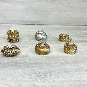 Può includere: Un assortimento di piccole scatole portagioie decorative in tonalità oro e argento. Le scatole presentano disegni ornati, alcuni con accenti di gioielli. Sono disposte su una superficie di legno grigio chiaro.
