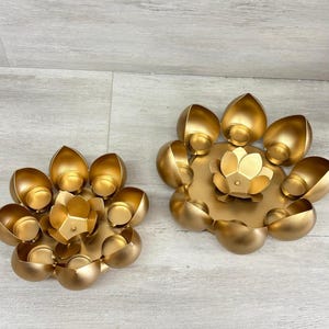 Könnte beinhalten: Zwei goldfarbene Lotusblumen-Kerzenhalter. Jeder Halter hat eine zentrale lotusförmige Basis, die von blütenblattförmigen Kerzenschalen umgeben ist. Die Halter sind aus Metall und werden auf einer hellen Holzoberfläche präsentiert.