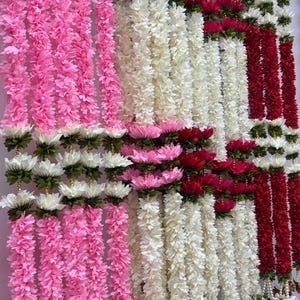Op de afbeelding: Een display van kleurrijke bloemenslingers. De slingers zijn in tinten roze, wit en rood, met witte en groene accenten. De slingers hangen verticaal, wat een levendig en feestelijk beeld cre&euml;ert.