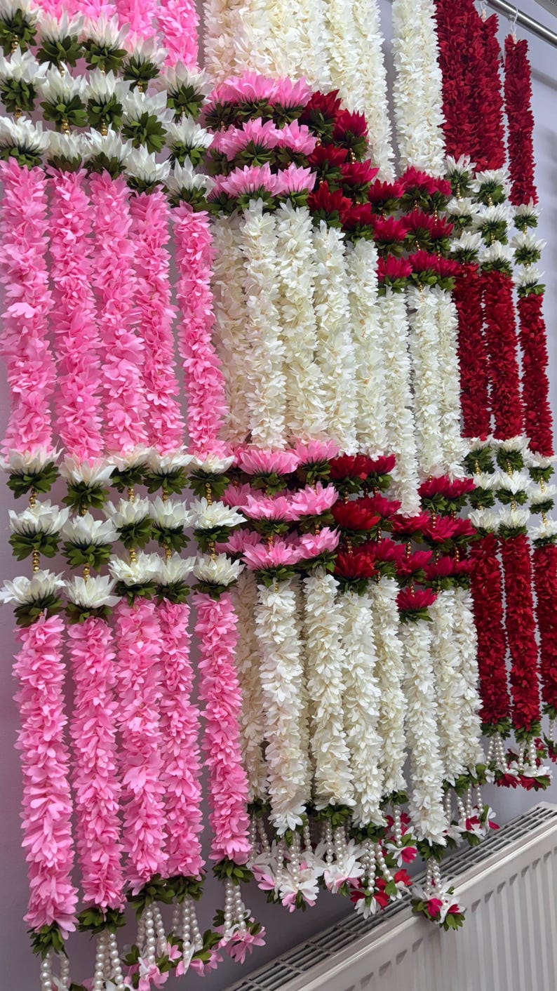 Op de afbeelding: Een display van kleurrijke bloemenslingers. De slingers zijn in tinten roze, wit en rood, met witte en groene accenten. De slingers hangen verticaal, wat een levendig en feestelijk beeld cre&euml;ert.