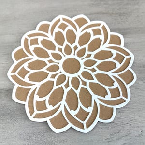 May include: A white paper cutout of a mandala design with a brown centre. The mandala has a circular shape and is made up of intricate patterns.