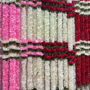 Op de afbeelding: Een display van kleurrijke bloemenslingers in roze, wit en rood. De slingers zijn verticaal gerangschikt, waardoor een levendige en decoratieve achtergrond ontstaat. De bloemen zijn gedetailleerd en lijken kunstmatig.