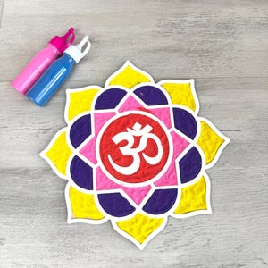 May include: A colourful mandala design with the symbol "Om" in the centre. The mandala is made of yellow, pink, purple, and red sand. Two bottles of paint are in the background.