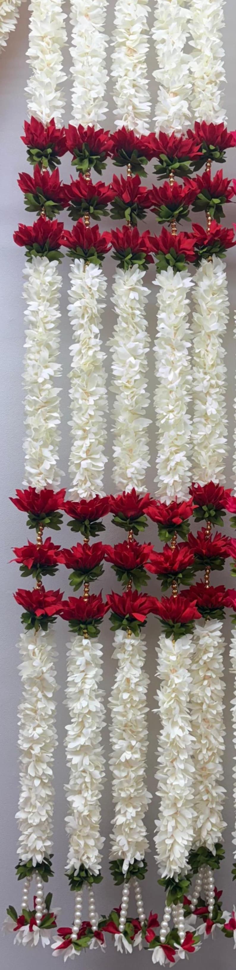 Op de afbeelding: Verticale weergave van bloemenslingers. De slingers zijn voornamelijk wit, met secties van levendige rode bloemen. De witte bloemen hebben een gelaagd bloemblaadjesontwerp. De rode bloemen zijn geaccentueerd met groene bladeren en gouden kralen.