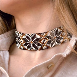 Könnte beinhalten: Ein Perlen-Choker-Halskette mit geometrischem Muster. Der Choker hat ein sich wiederholendes Design aus weißen, braunen und goldenen Perlen. Die Halskette wird um den Hals getragen und ist auf einem hellen Hemd sichtbar.