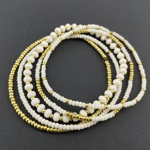 May include: A collection of beaded bracelets. The bracelets feature a combination of white and gold beads. The white beads are larger and more rounded, while the gold beads are smaller and more numerous. The bracelets are arranged in a circular pattern.