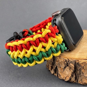 May include: A black smartwatch with a colorful, woven band in red, yellow, and green. The band is made of braided cord and is attached to the watch. The watch rests on a wooden surface.