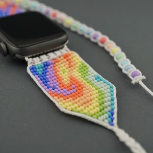Op de afbeelding: Een regenboogkleurig kralenhorlogebandje met een wit koord en een zwarte Apple Watch.
