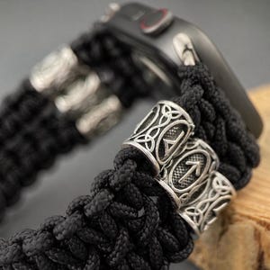 May include: Black braided watch strap with silver metal detailing. The strap has decorative metal beads with engraved symbols. The watch is black and partially visible, resting on a wooden surface.