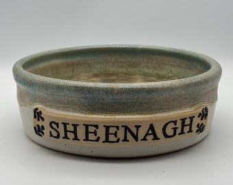 May include: A round ceramic bowl with a glazed finish in shades of blue, green, and cream. The exterior features a raised rectangular plaque with the name SHEENAGH embossed in black capital letters, flanked by small black leaf-like decorative motifs.