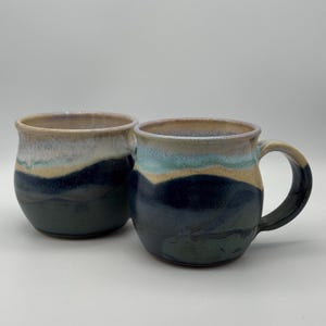 May include: Two handcrafted ceramic mugs with a distinctive layered design. The mugs have a dark blue base, with bands of tan, light blue, and a light brown rim. One mug has a handle, the other does not. Ideal for tea or coffee.