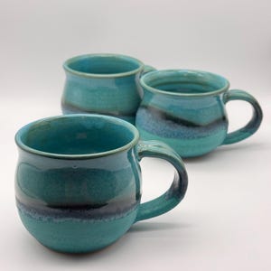 May include: Three teal and blue ceramic mugs. Each mug has a rounded body, a curved handle, and a dark blue band around the middle. The mugs are glazed with a glossy finish. The mugs are arranged on a white background.