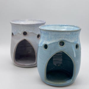 May include: Two ceramic oil warmers in soft blue and lavender hues. Each warmer has a rounded shape with a large arched opening and three small circular holes. The warmers are glazed with a glossy finish.