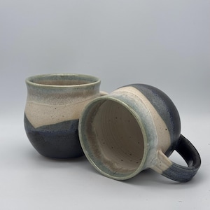 May include: Two handmade ceramic mugs. Each mug has a rounded body with a handle and a unique colour scheme. The colour palette includes shades of blue, beige, and grey, creating a layered effect. The mugs are designed for beverages.